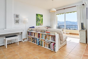A living room with a sofa, a bookshelf, a piano, and a balcony with a view.