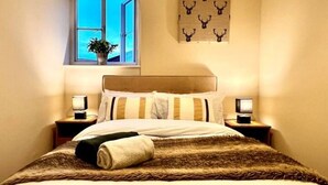 A neatly made bed with a striped pillow, a fur throw, and a towel on the side. There are two bedside lamps on wooden nightstands. A window with a view of the sky and a plant on the sill is visible.