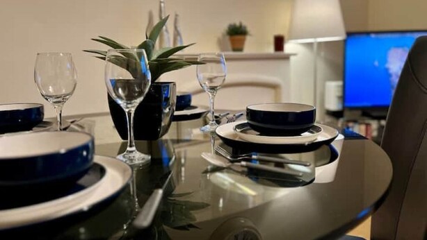 A dining table set with plates, bowls, and wine glasses, with a potted plant and a laptop in the background.