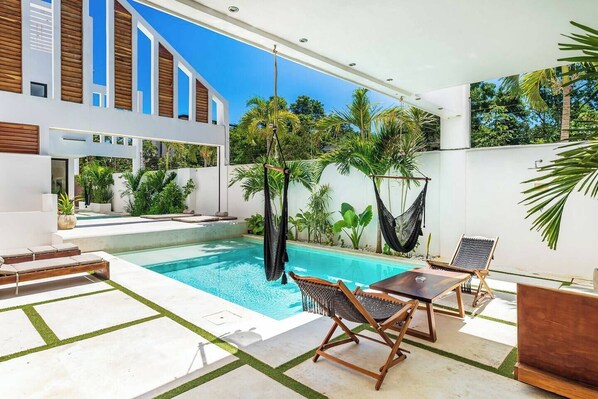 Un espace piscine extérieur moderne, avec une piscine à l’eau d’un bleu limpide, des chaises longues en bois, un hamac et une végétation tropicale luxuriante.