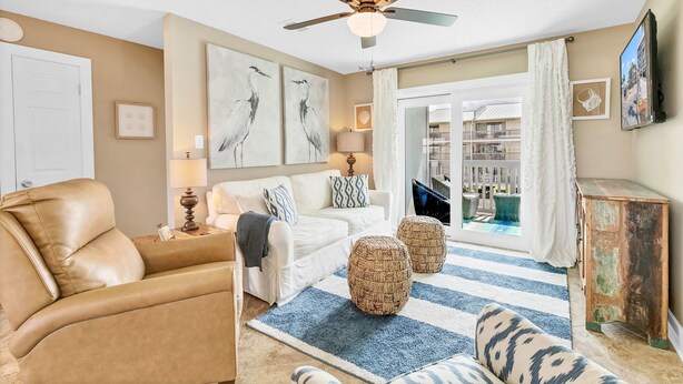 A living room with a beige sofa, a white couch, and a ceiling fan. There are two wicker ottomans on a striped rug, a lamp, and a television on a wooden cabinet. - Orange Beach Oasis - Old River Front with 1 minute walk to beach! (Orange Beach)