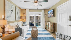 A living room with a sofa, ottoman, and a ceiling fan. There is a TV mounted on the wall and a door leading to another room. - Orange Beach Oasis - Old River Front with 1 minute walk to beach! (Orange Beach)