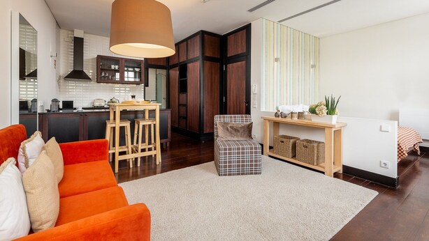 A modern living room with a sofa, a coffee table, and a dining area with a table and chairs. There is a kitchen area with cabinets and a hanging light fixture.