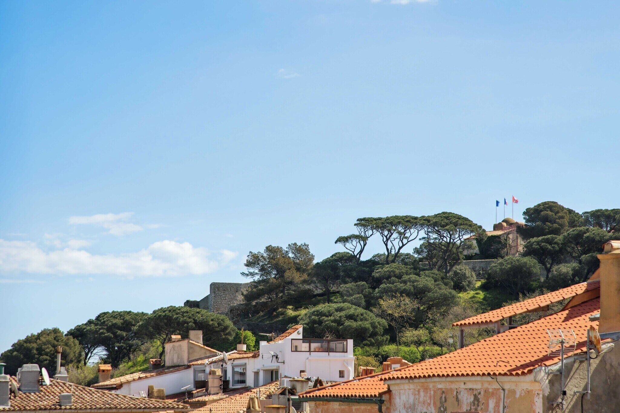 Une ville située sur une colline, avec des bâtiments aux toits de tuiles rouges, un imposant mur de pierre et une végétation dense.