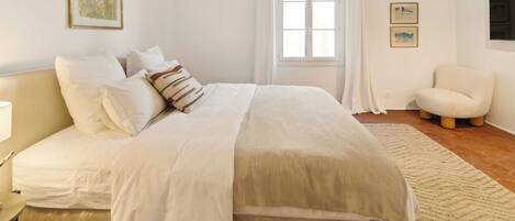 Ein Schlafzimmer mit einem Bett, einem Sessel, einem Fenster mit Vorhängen und Bildern an den Wänden.