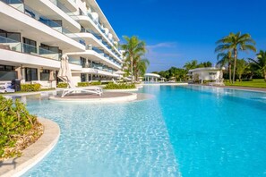 A modern hotel with a large swimming pool, multiple balconies, and palm trees. - Beautiful Romantic Condo, Balcony with Jacuzzi and Spectacular Golf Course Views (Punta Cana)