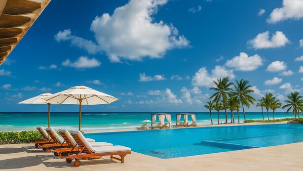 Zona junto a la piscina con sillones de descanso y sombrillas, cielo azul despejado y vista al océano.