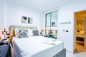 A modern bedroom with a large bed, bedside tables, a nightstand, a mirror, and a view into a bathroom. - Beautiful Romantic Condo, Balcony with Jacuzzi and Spectacular Golf Course Views (Punta Cana)
