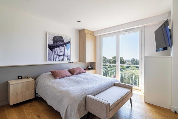 Une chambre moderne avec un grand lit, un banc, une table de chevet, un téléviseur fixé au mur et une grande fenêtre donnant sur un beau paysage.