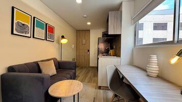 En moderne lejlighed med en mørkegrå sofa, et lille rundt bord, et spiseområde med en hvid stol, et køkken med et træskab og et stort vindue med udsigt til bygninger.