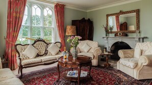 A traditional living room with a fireplace, two sofas, a coffee table, and a large window with curtains.