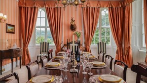 A dining room with a long table set for a meal, featuring a chandelier, curtains, and a view of greenery outside.