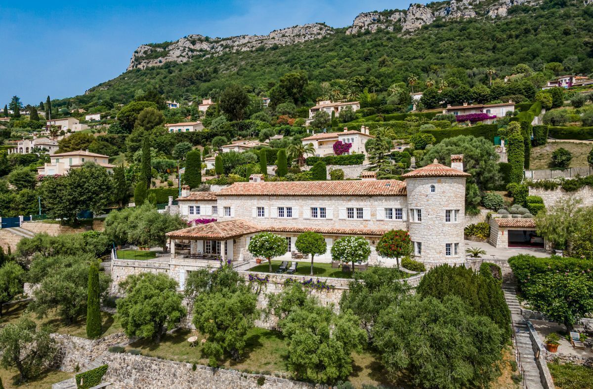 Une villa située sur une colline, avec une façade en pierre, un toit en tuiles de terre cuite et un jardin agrémenté d’arbres et d’arbustes adultes.