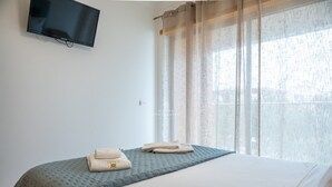 Ett hotellrum med en säng, en platt-TV som är monterad på väggen och ett fönster med tunna, genomskinliga gardiner.