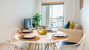 En matplats med ett runt bord för fyra personer, omgiven av vita stolar. Ett stort fönster med gardiner syns i bakgrunden, och en soffa med kuddar står placerad nära fönstret.