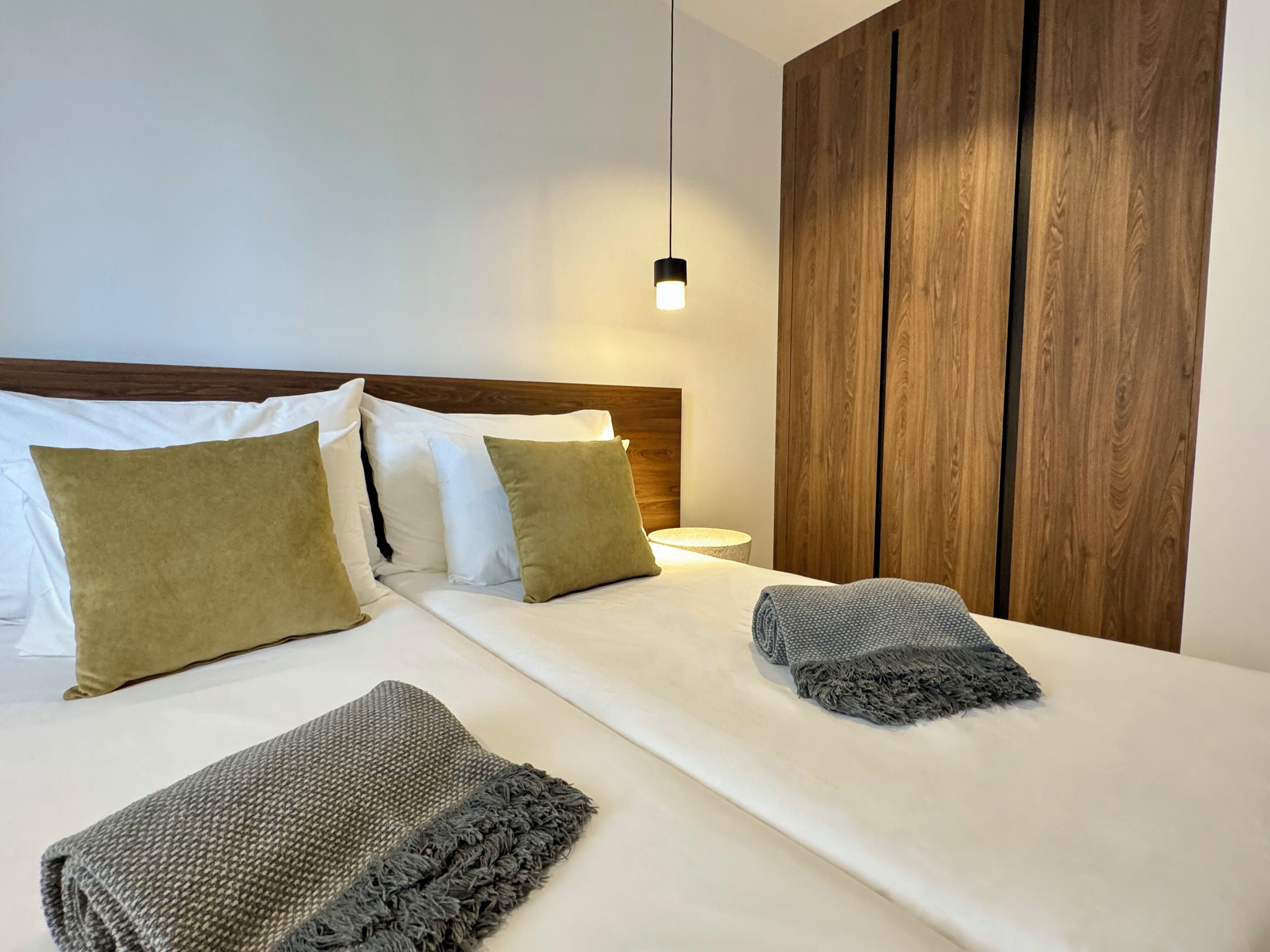Een netjes opgemaakt bed met witte en bruine kussens, een grijze plaid en een geweven deken. Er is een houten garderobe en een bedlampje.