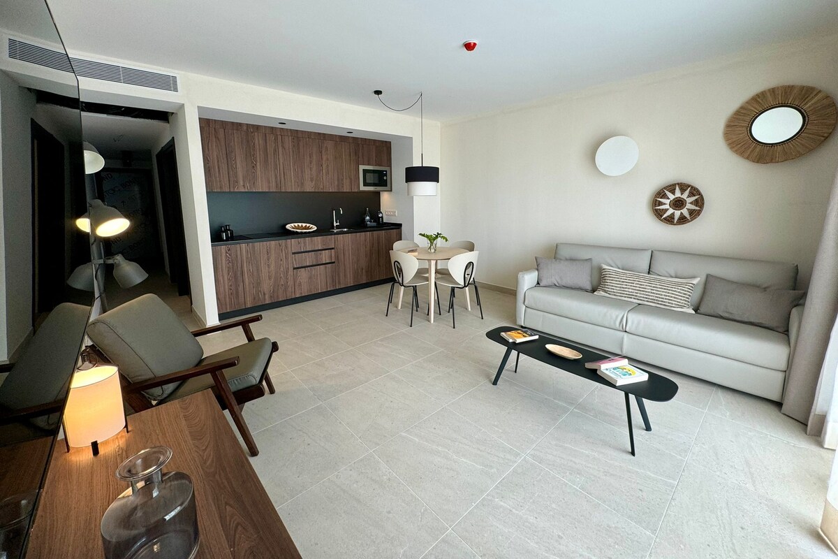 A modern living room with a sofa, coffee table, and chairs, a kitchen area with wooden cabinets, and a dining table with chairs.