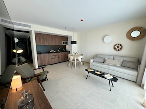 Ein modernes Wohnzimmer mit einer Couch, einem Couchtisch und Sesseln. Im Hintergrund ist eine Küchenzeile mit Holzschränken und einem Esstisch zu sehen.
