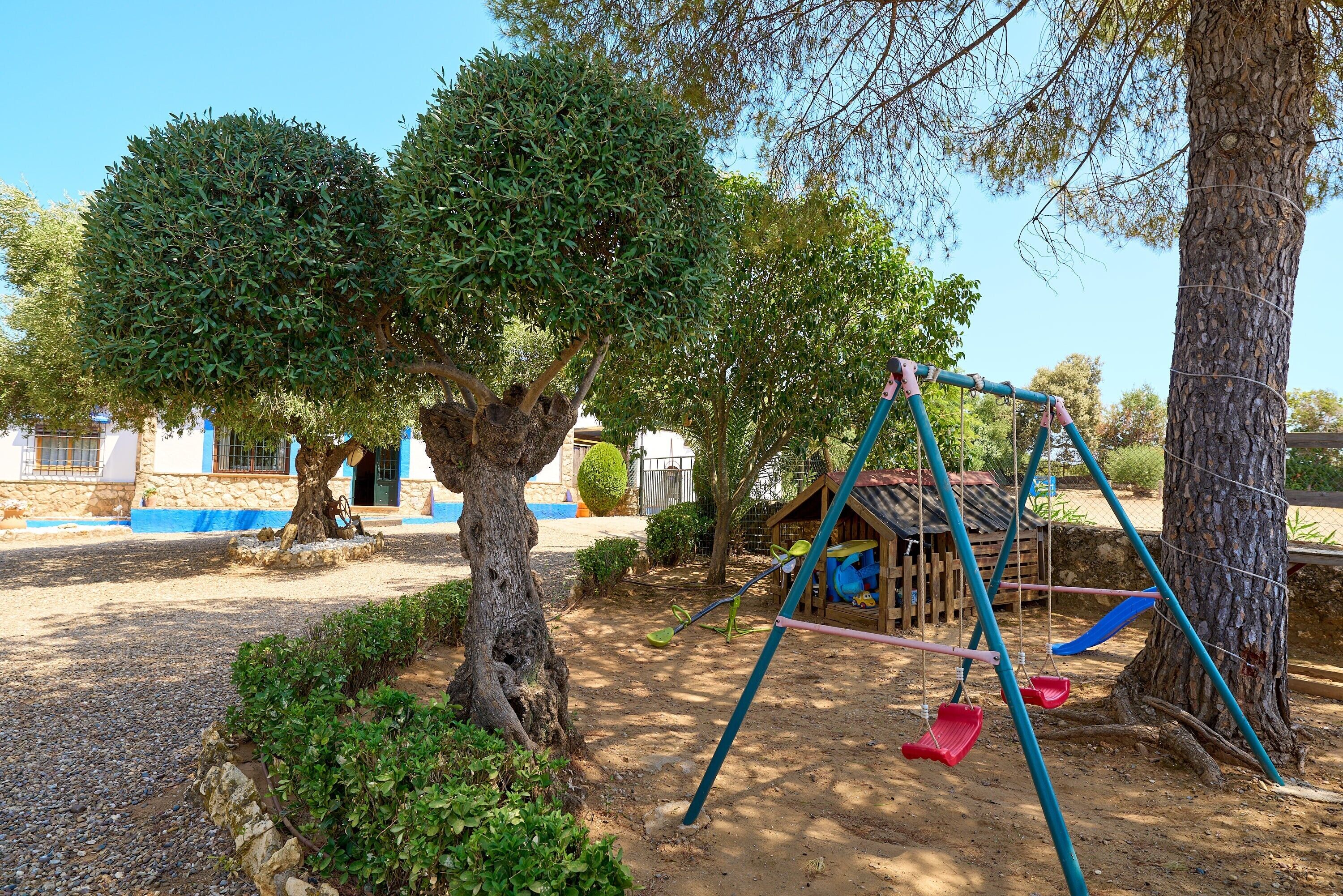 Ein Spielplatz mit Schaukeln und Rutsche, umgeben von Bäumen und einem Gebäude mit blau-weißer Fassade.