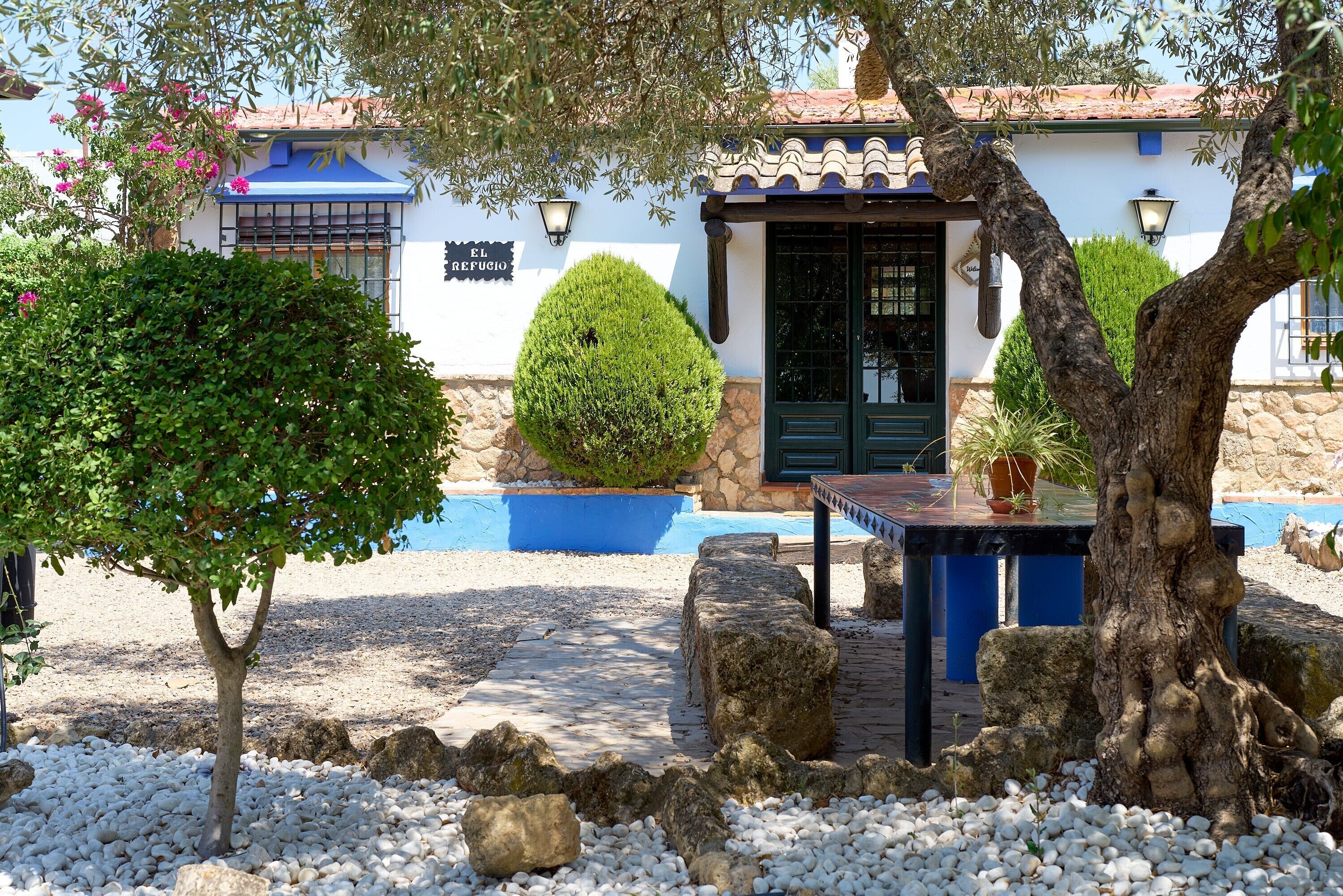 Un edificio con un letrero que dice 'El Refugio', un patio con una mesa y sillas, y un pequeño árbol.