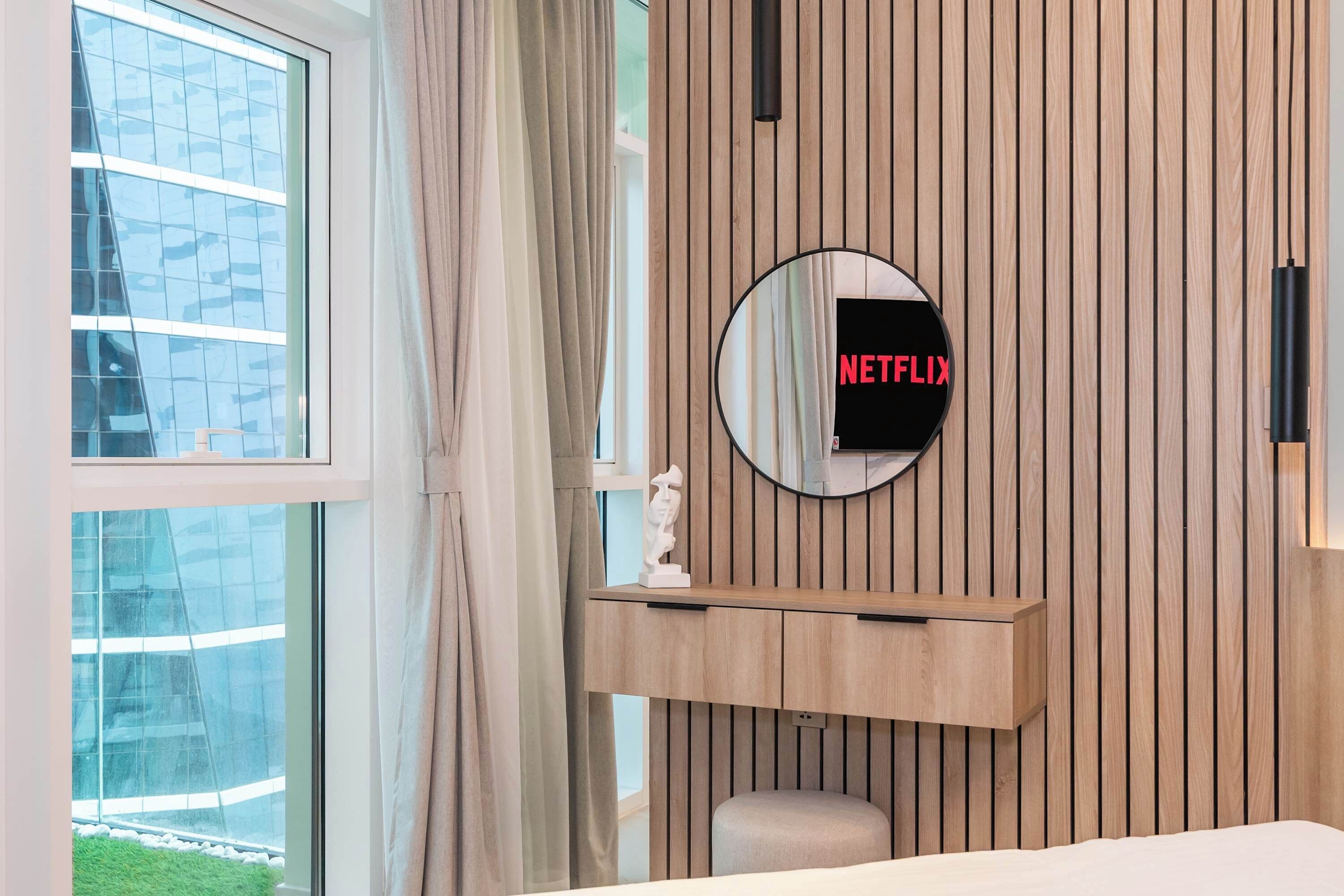 Moderni hotellihuone, jossa on suuri ikkuna, puupanelilla verhoiltu seinä, pyöreä peili, jossa on Netflix-logo, pieni patsas ja yöpöytä.