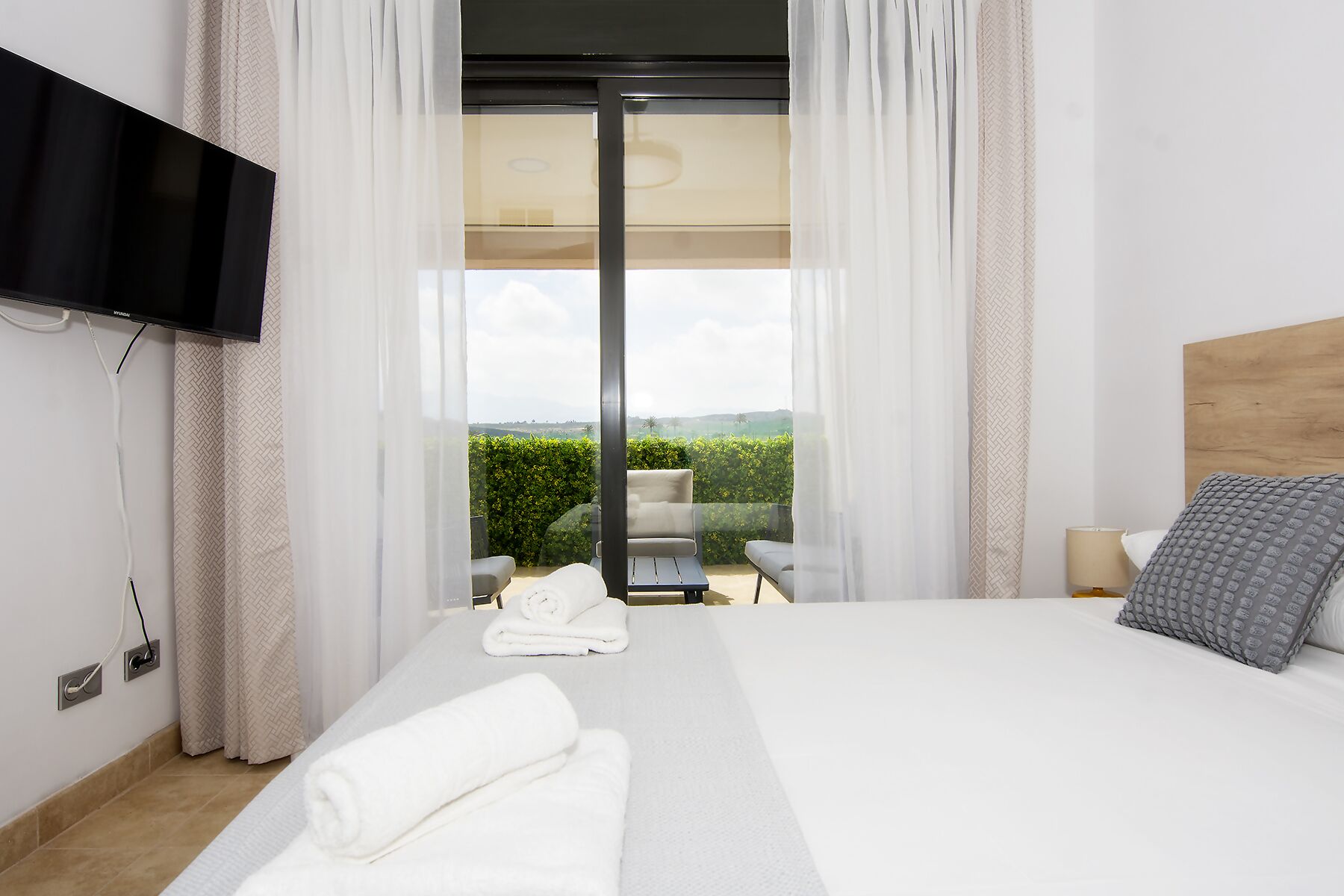 A modern hotel room with a large bed, a flat-screen TV mounted on the wall, a balcony with a chair, and a view of the countryside.