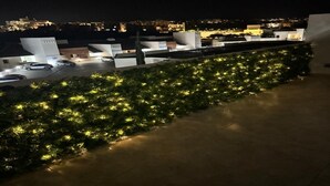 Un toit-terrasse bordé d’une rangée d’arbres illuminés par des lumières, un parking avec des voitures et des bâtiments en arrière-plan.