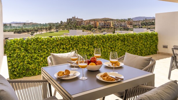 Une table à manger dressée avec des croissants, des fruits et du jus d’orange, donnant sur un paysage urbain avec une haie au premier plan.