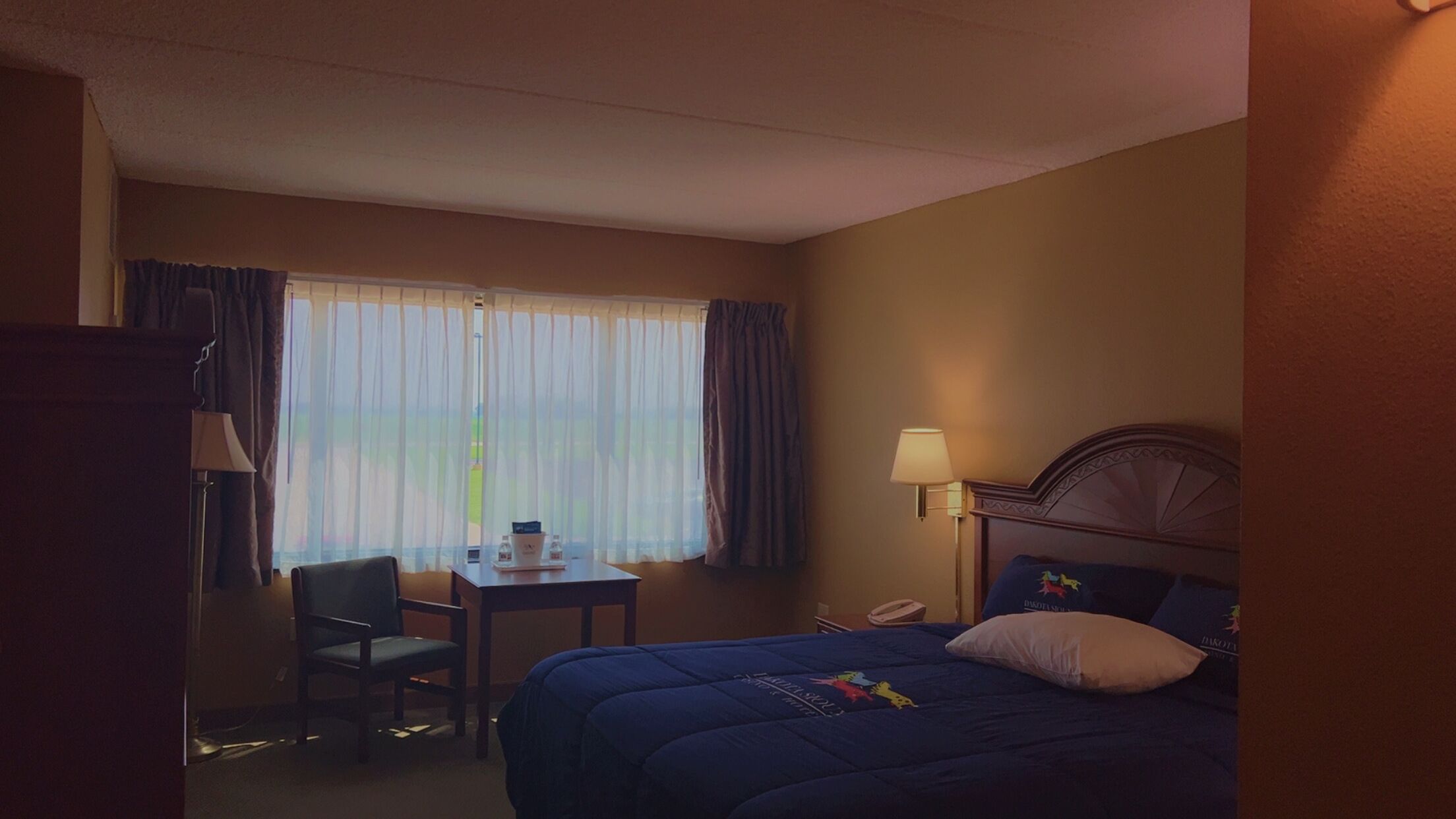 A hotel room with a bed, a desk, a chair, a window with curtains, and a lamp.