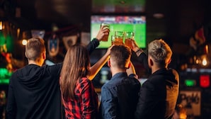 Un grupo de personas brindando con bebidas en un bar, mientras se ve un evento deportivo en la televisión.