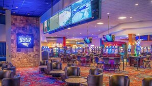 Un casino con múltiples mesas de juego y máquinas tragamonedas, asientos de cuero y una pantalla grande que muestra eventos deportivos.