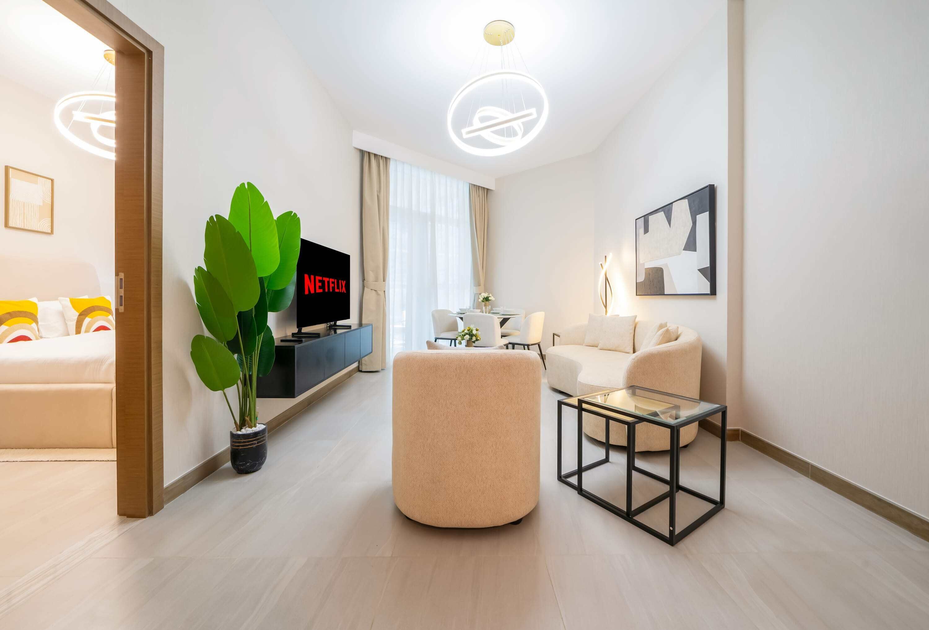 Una habitación de hotel moderna con sofá, reposapiés y una mesa de centro de vidrio. Se observa un televisor de pantalla plana montado en la pared y una planta en maceta.