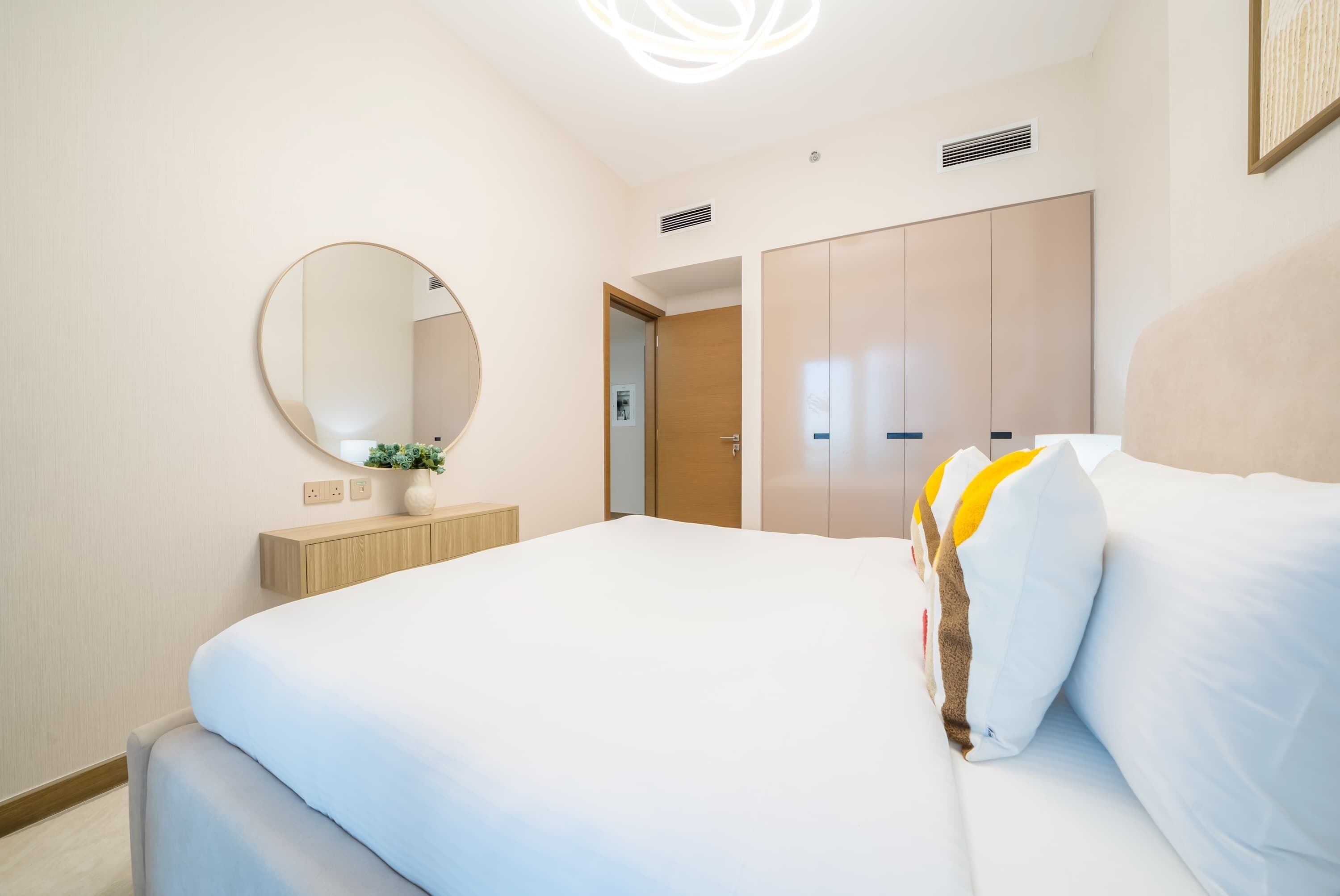 A modern hotel room with a large bed, a round mirror, a wardrobe, and a small bedside table with a vase.