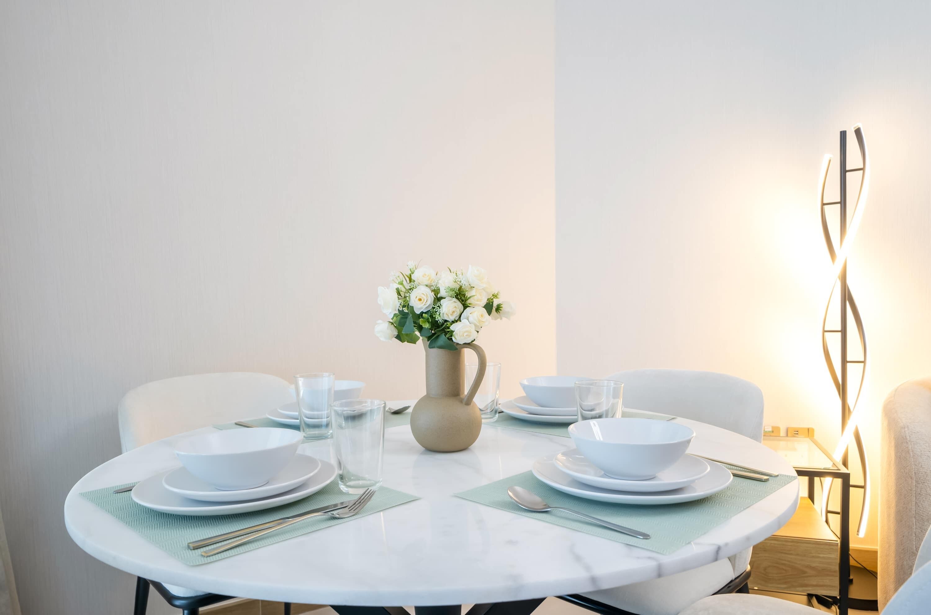A dining table set with white plates, bowls, and cutlery, a vase with white flowers, and a modern light fixture.