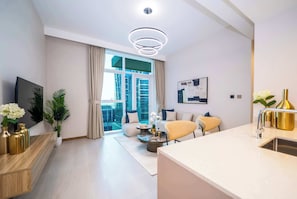 Un salon moderne doté d’une grande fenêtre, d’un téléviseur à écran plat, d’un canapé, d’une table basse et d’un coin cuisine avec un évier et des accessoires dorés.