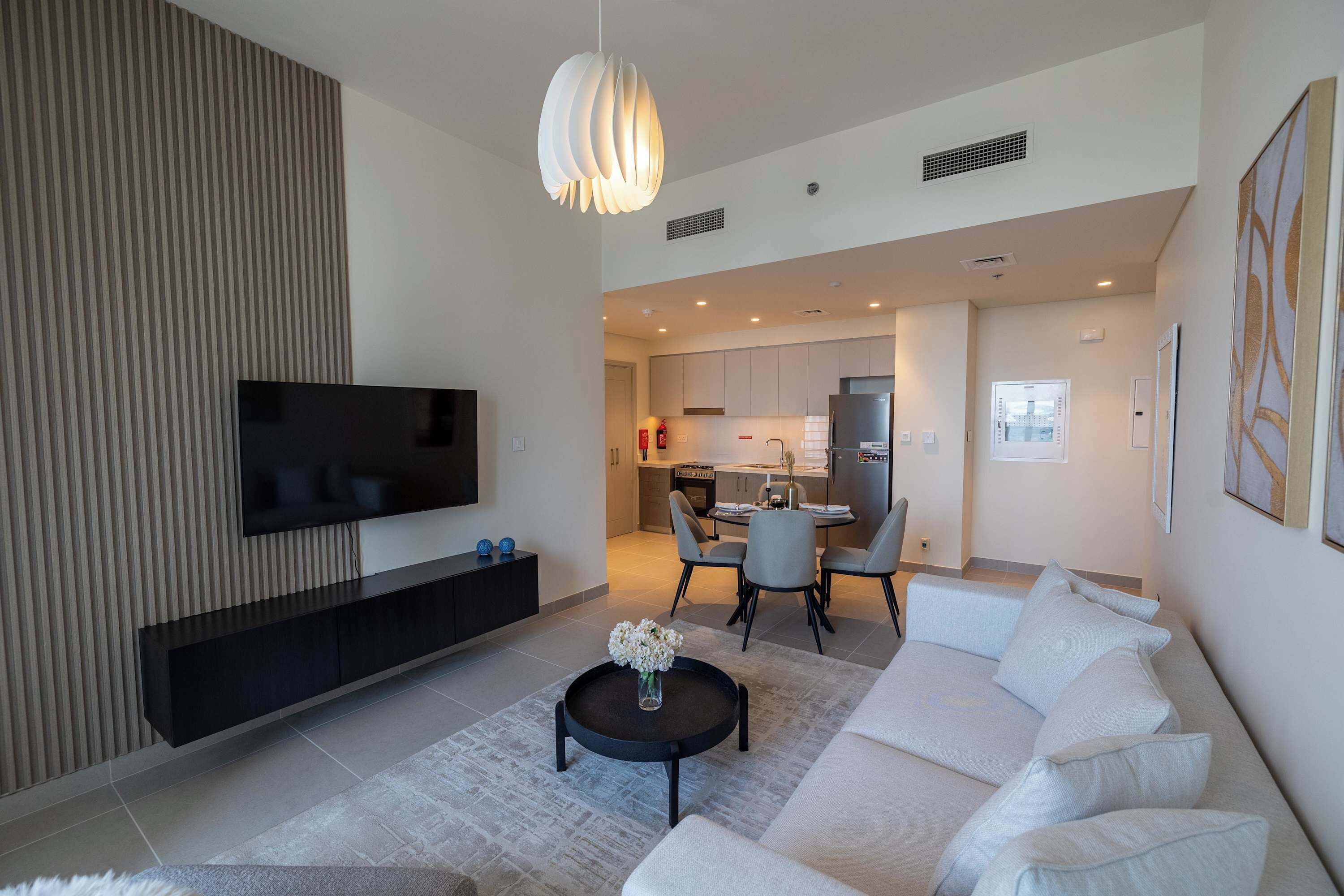 Ein modernes Wohnzimmer mit einem Flachbildfernseher, einem schwarzen Couchtisch mit Blumenvase, einem weißen Sofa und einem Essbereich mit Tisch und Stühlen.