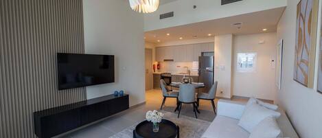 A modern living room with a flat-screen TV, a black coffee table with a vase of flowers, a white sofa, and a dining area with a table and chairs.