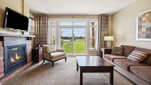 A living room with a fireplace, a sofa, a chair, a TV, and a view of a golf course through large glass doors. - Blue Sky Retreat at Rivergrass (The Blue Mountains)