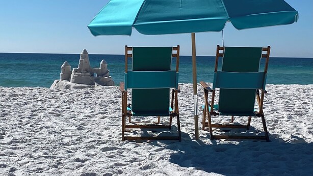 Two beach chairs under a blue umbrella, a sandy beach, and a sandcastle in the background.