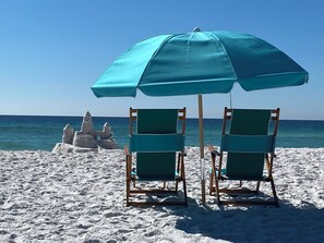 Två strandstolar under ett blått parasoll, en sandstrand och ett sandslott i bakgrunden.