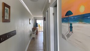 A hallway with a painting of a sunset beach scene, a door, and a dining area in the background.