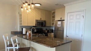 A kitchen with a central island, white bar stools, a stainless steel refrigerator, a microwave, and a hanging light fixture.