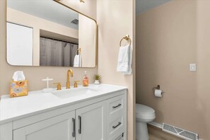 Une salle de bain avec un lavabo blanc, un robinet doré et un miroir au-dessus. On y trouve des toilettes, une serviette accrochée au mur et une grille d’aération au sol.