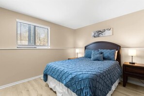 Une chambre à coucher comprenant un lit, deux tables de chevet, une fenêtre avec des stores et une œuvre d’art accrochée au mur.