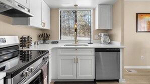 A modern kitchen with white cabinetry, a stainless steel stove, a dishwasher, and a window with blinds. - NEW Corp/Bus Townhome New Hudson (New Hudson)