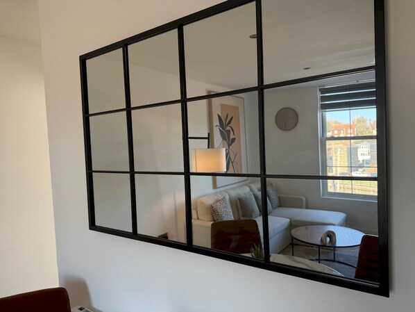 A room with a large mirror reflecting a living area, a sofa, a lamp, and a view of buildings outside.