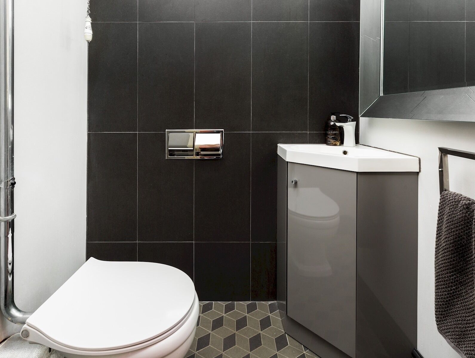 Un baño moderno con inodoro blanco, paredes de baldosas oscuras, lavamanos blanco y un espejo sobre el lavamanos.