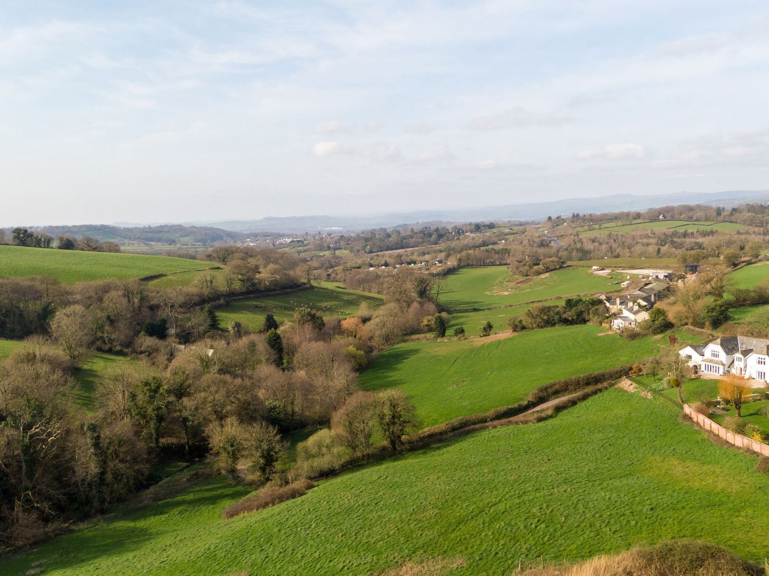 Paisagem rural com campos verdes ondulantes, árvores dispersas e algumas casas.