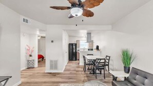 A modern living room with a ceiling fan, a dining area with a table and chairs, and a kitchen in the background.