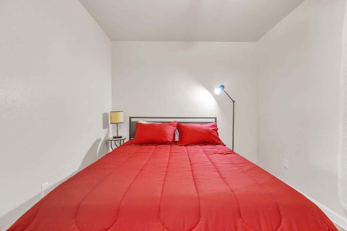 A single bed with a red bedspread and two pillows, a bedside lamp, and a small table with a lamp.